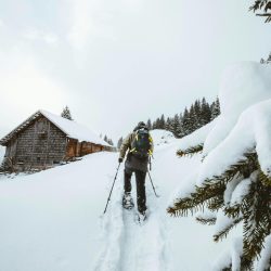 A hiker traverses a snowy mountain path towards a cabin, surrounded by winter scenery.