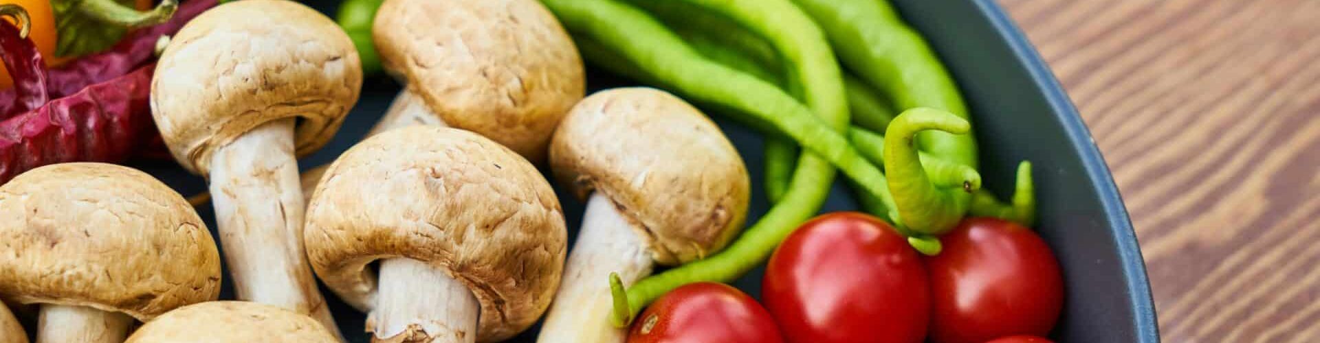 A close-up of various fresh vegetables and mushrooms in a pan on a wooden surface.