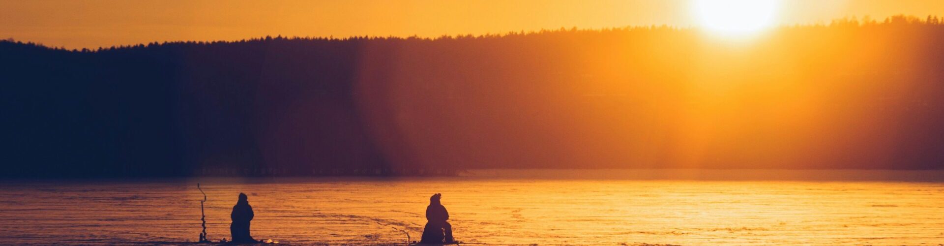 Silhouetted figures ice fishing on a frozen lake during a vibrant sunset in Lahti, Finland.