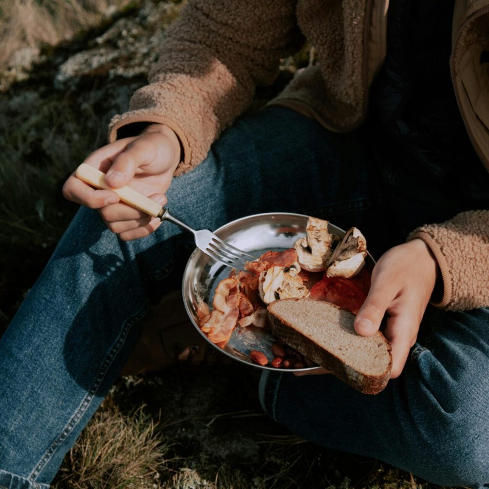 A person enjoys a rustic outdoor meal with bread, meat, and mushrooms on a metal plate.