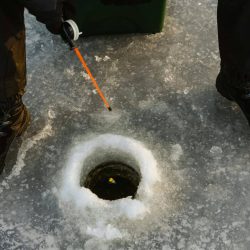 Person ice fishing on frozen lake surface with fishing rod and winter boots.