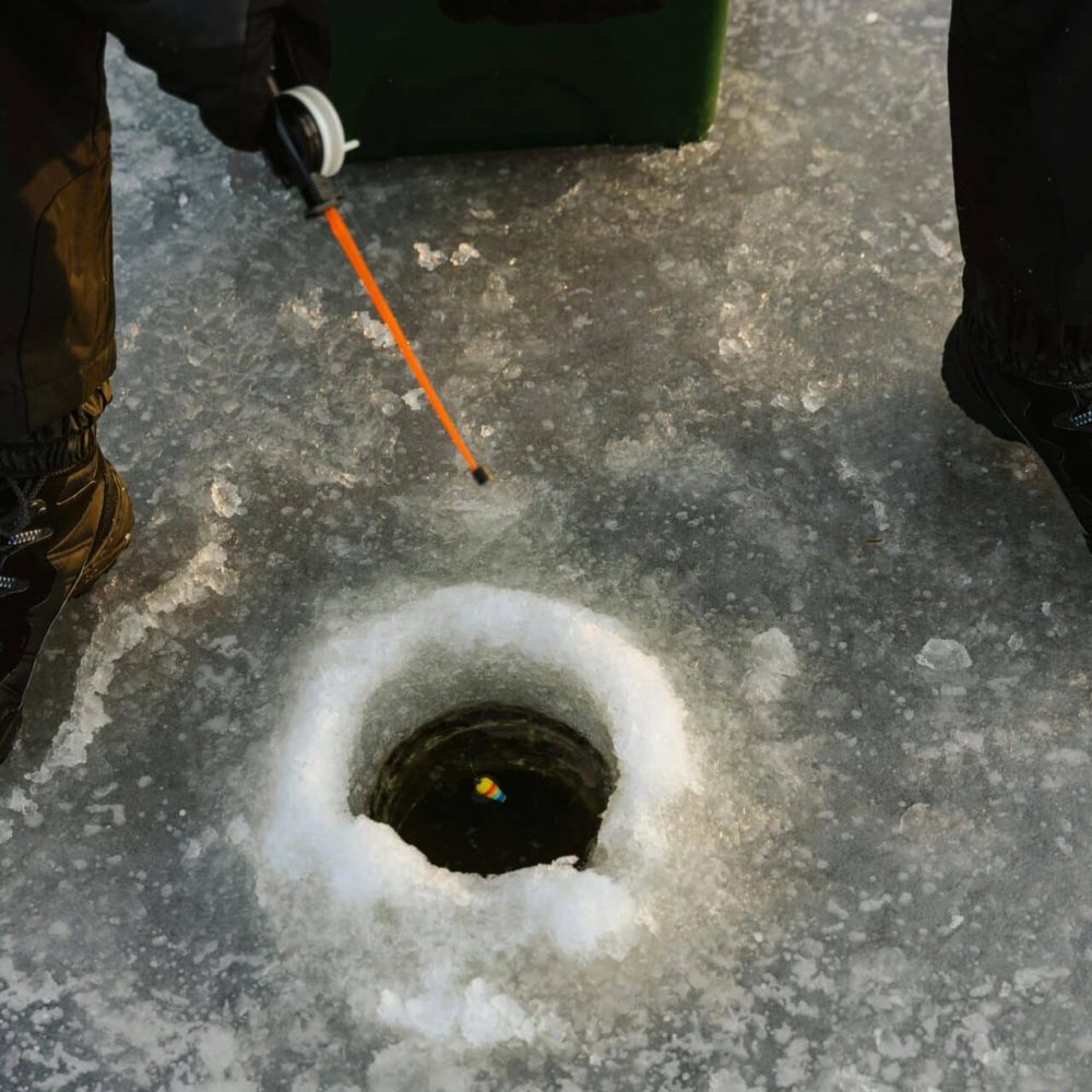 Person ice fishing on frozen lake surface with fishing rod and winter boots.
